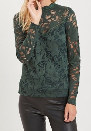 Top lungo a maniche lunghe verde in pizzo con un motivo floreale, colletto alto e maniche trasparenti, abbinato a pantaloni neri aderenti.