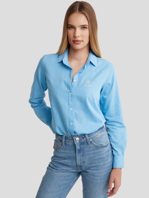 Hellblaues Langarmhemd mit vertikalen weißen Streifen, Button-Down-Kragen und gesticktem Logo auf der Brust, kombiniert mit hoch taillierten Jeans.