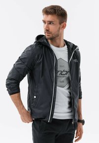 Sort zip-up jakke med hætte, der har elastiske manchetter og en glat tekstur; lagdelt over en grå grafisk t-shirt.