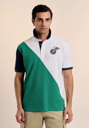Hombre vistiendo una camisa polo verde y blanca con cuello y mangas azul marino, pantalones cargo beige, de pie frente a un fondo liso.