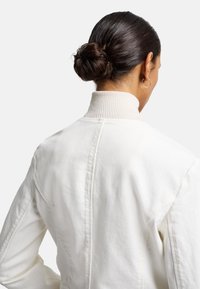 Witte jas met een hoge geribbelde kraag en een textuurstof. De achterkant heeft naden en een algehele slanke snit. Haar in een knot gestyled.