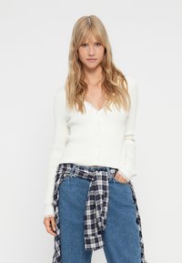 Λευκή ριγέ μακρυμάνικη κάρντιγαν με δαντελένια περιθώρια στον λαιμό, κουμπί κλεισίματος, σε συνδυασμό με μπλε τζιν και σκούρο καρό πουκάμισο δεμένο στη μέση.