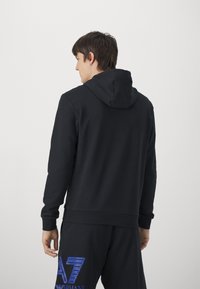 Felpa nera con cappuccio e maniche lunghe. Presenta una texture liscia e orli a coste. Abbinata a pantaloni della tuta neri con un logo blu.