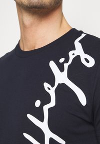 Maglietta di cotone blu navy con una grande grafica astratta bianca sul lato sinistro, maniche corte e scollatura rotonda.