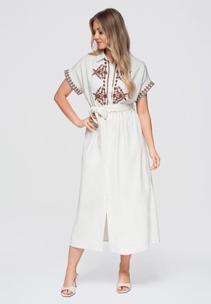 Femme portant une robe midi blanche avec broderie florale marron, manches courtes, boutons sur le devant, ceinture nouée à la taille et talons ouverts beige.