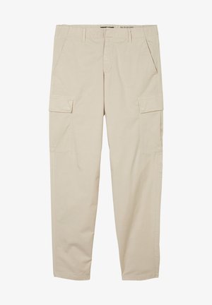 Person trägt olivgrüne Jacke, dunkelgraues T-Shirt, beige Cargohose, schwarzen Gürtel und weiße Sneakers mit braunen Sohlen, steht vor einer weißen Wand.
