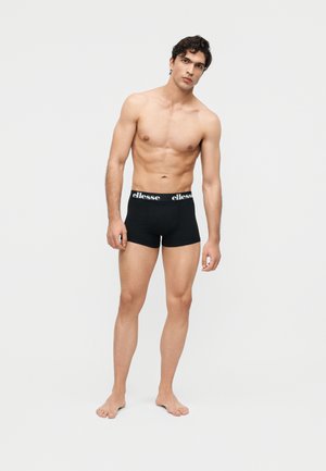 Young man standing barefoot wearing black Ellesse boxer briefs against a plain light background.