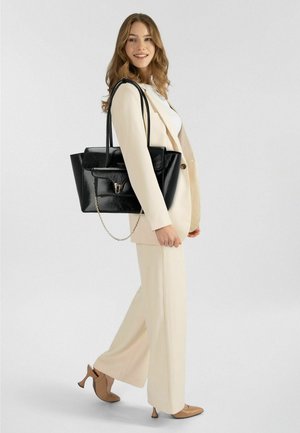 Zwarte lederen totebag met een voorvak, kettingaccent en dubbele schouderbanden. Model draagt een crèmekleurig pak en beige hakken.