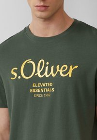 Grünes Baumwoll-T-Shirt mit kurzen Ärmeln, das ein großes goldfarbenes "s.Oliver"-Logo und Text darunter zeigt, mit Rundhalsausschnitt und glatter Textur.