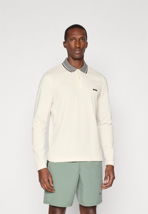 Long-sleeve polo shirt in cream with a gray collar, featuring a button placket and a black logo on the chest, paired with green shorts.