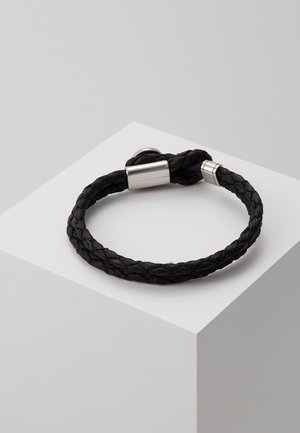 Schwarzes geflochtenes Lederarmband mit silberner Magnetverschluss, dargestellt auf einem weißen würfelförmigen Podest vor grauem Hintergrund.