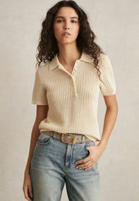 Polo shirt a maglia di colore crema con maniche corte e colletto, dotata di una patta con bottoni e una vestibilità morbida, abbinata a jeans blu.