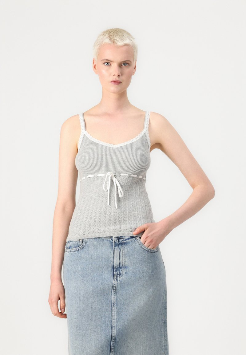 Szary, prążkowany top na ramiączkach z białą koronką, z przodu z detalem w postaci sznureczka. Modelka nosi go z jasnoniebieską spódnicą dżinsową.