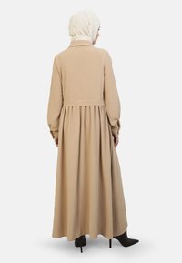 Maxikjole i beige stof med lange ærmer og rund krave. Har en figursyet talje og en svajende nederdel. Båret med en hvid hijab.