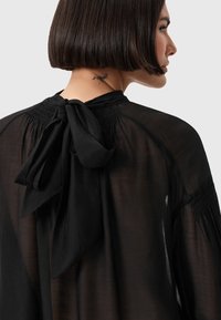Blusa nera trasparente con chiusura a fiocco sul retro, dettagli arricciati lungo lo scollo e texture liscia.