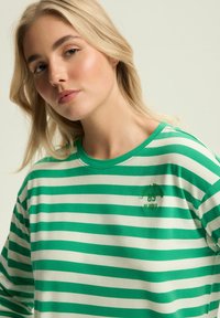 Zielona i biała w paski bluzka z długim rękawem z miękkiego materiału, z okrągłym dekoltem i małym haftem na klatce piersiowej.