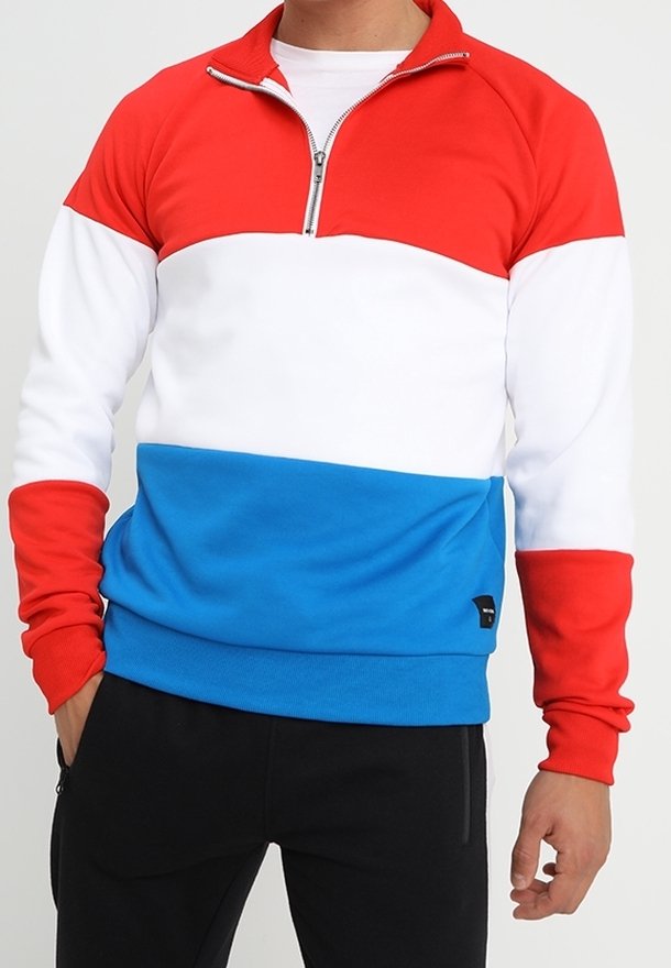 Uomo che indossa un pullover a maniche lunghe con blocchi di colore rosso, bianco e blu e una zip frontale, abbinato a pantaloni neri.