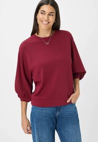 Bordeaux oversized pullover sweatshirt met driekwartballonmouwen, geribbelde onderrand en een zachte textuur. Jeans zichtbaar bij de taille.