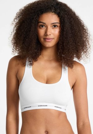 White sports bra featuring a scoop neckline and wide straps, with a branded elastic band displaying "GUESSJEANS." Smooth, stretchy fabric.