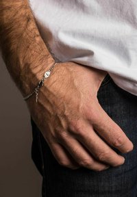 Ezüst lánc karkötő virág medállal, egy csuklón viselve. Texturált lánccal és homár kapoccsal rendelkezik, egy texturált sötét háttér előtt.