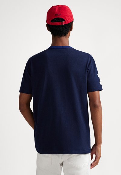 Tmavě modré bavlněné tričko s krátkými rukávy, s kontrastním límcem a bílými čísly na levém rukávu. Doplněno červenou čepicí.