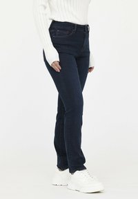 Jeans de tiro alto color azul oscuro, confeccionados en denim, con un corte ajustado y bolsillos frontales, combinados con una blusa de punto blanca y zapatillas blancas.