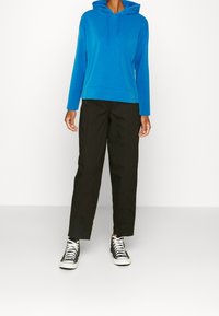 Modrá bavlněná mikina s kapucí na stažení, doplněná černými zúženými kalhotami a černými vysokými teniskami, zobrazena na jednoduchém pozadí.