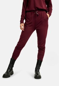 Bordové tepláky s jemnou textúrou, elastickým pásom so zaväzovaním a zužujúcimi sa nohavicami; skombinované s čiernymi členkovými čižmami.