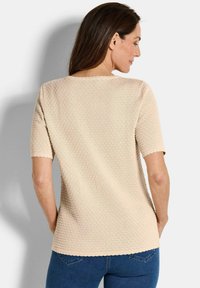 Vrouw in een beige gestructureerde top met korte mouwen en gegolfde randen, gezien van achter, gecombineerd met blauwe jeans.