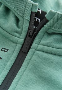 Grüne Zip-Hoodie aus strukturiertem Stoff mit schwarzem Reißverschluss und Logoakzenten. Glatte Nähte entlang der Säume und des Kragens.
