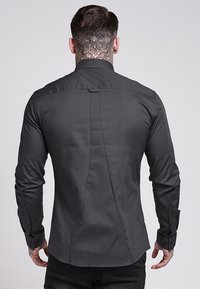 Camisa de botones de tela gris oscuro, con una costura en la parte posterior y puños con botones. Textura suave, ajuste entallado y mangas largas.