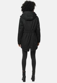 Black parka with an adjustable hood, elastic waist, and a longer back. Features a smooth texture and snap-button closures. Paired with dark jeans and boots.
