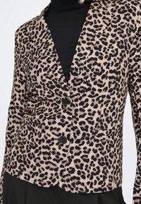 Femme portant un blazer beige et noir à imprimé léopard avec des boutons noirs sur un col roulé noir et un pantalon noir.
