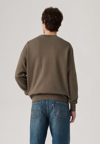 Sudadera de color verde oliva con puños y dobladillo acanalados, combinada con jeans de color azul claro, ligeramente desgastados, que cuentan con bolsillos traseros y costuras.