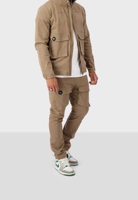 Beige bomber jacket dengan dua kantong dada besar, dipadukan dengan celana cargo serasi yang memiliki kantong samping. Alas kaki berupa sneakers putih dan hijau.