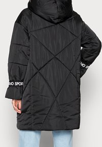 Abrigo acolchado negro con capucha, que presenta un patrón acolchado en forma de diamante y detalles contrastantes "JUJO SPORT" en blanco en los puños. Textura lisa.