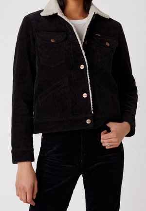 Personne portant une veste en velours côtelé noir avec un col en sherpa blanc sur une chemise blanche, associée à un pantalon en velours côtelé noir assorti.