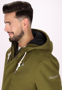 Giacca verde oliva con chiusura a zip, cordini bianchi, fodera interna nera e dettaglio del logo sulla manica; presenta un cappuccio e un tessuto texture.