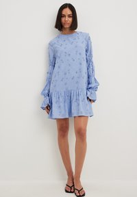 Svetlo modra obleka iz teksturirane tkanine, ki ima naborkane dolge rokave in večplastni rob, okrašen z majhnimi vezenimi rožicami.