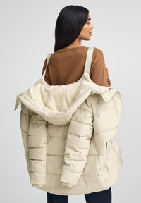 Beżowa kurtka puchowa z poziomym pikowaniem, oversize'owym krojem i wyściółką wewnętrzną. Posiada regulowane paski z logo oraz kaptur.