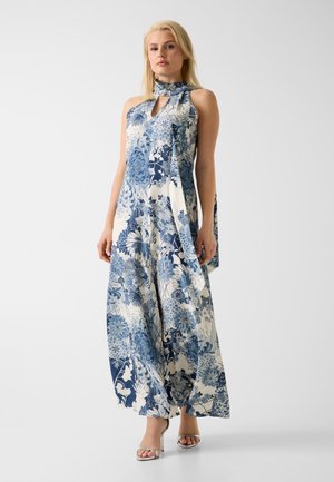 Donna bionda che indossa una tuta senza maniche con collo all'americana con stampa floreale blu e bianca, abbinata a sandali argentati con tacco alto.