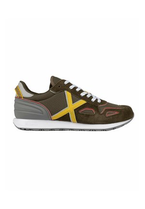Zapatilla de ante verde oliva con acentos amarillos. Cuenta con cordones blancos, talón gris y paneles laterales texturizados. Suela de goma blanca y gris.