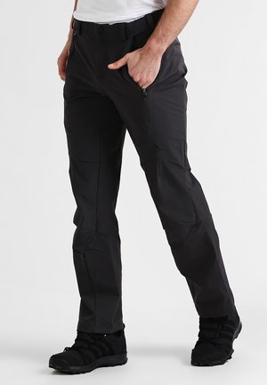 Mężczyzna w czarnych spodniach Adidas z zapinanymi na zamek bocznymi kieszeniami i czarnych butach sportowych, stojący na tle jednolitego białego tła.