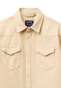 Chemise beige à boutons-pression, col pointu, deux poches poitrine avec rabats, et une étiquette noire "Jean's West" à l'intérieur du col.