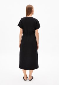Schwarzes, knielanges Kleid aus glattem Stoff. Verfügt über kurze Ärmel, einen elastischen Bund und eine gerade Silhouette. Getragen mit flachen Sandalen.