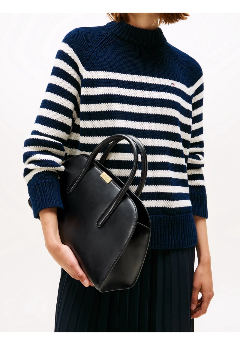 Personne portant un pull rayé bleu marine et blanc tenant un sac à main en cuir noir avec des anses doubles et une étiquette dorée.