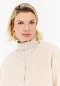 Blonde vrouw met blauwe ogen, gekleed in een crèmekleurige turtleneck en een gemêleerde fleecejack, die iets opzij kijkt tegen een witte achtergrond.