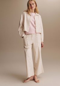 Blonde vrouw staat in een lichtbeige cropped jack, bijpassende wijde pijpen broek, lichtroze overhemd en beige platte sandalen tegen een neutrale achtergrond.