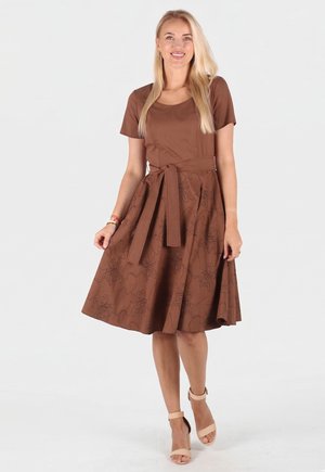 Blonde Frau lächelt, trägt ein braunes Kleid mit kurzen Ärmeln, floralem Muster und gebundenem Gürtel, kombiniert mit beigen Sandalen mit hohen Absätzen.