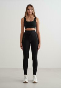Leggings negre cu talie înaltă și şnur, asortate cu un top fit negru scurt; ambele realizate dintr-un material neted, purtate cu adidași albi.